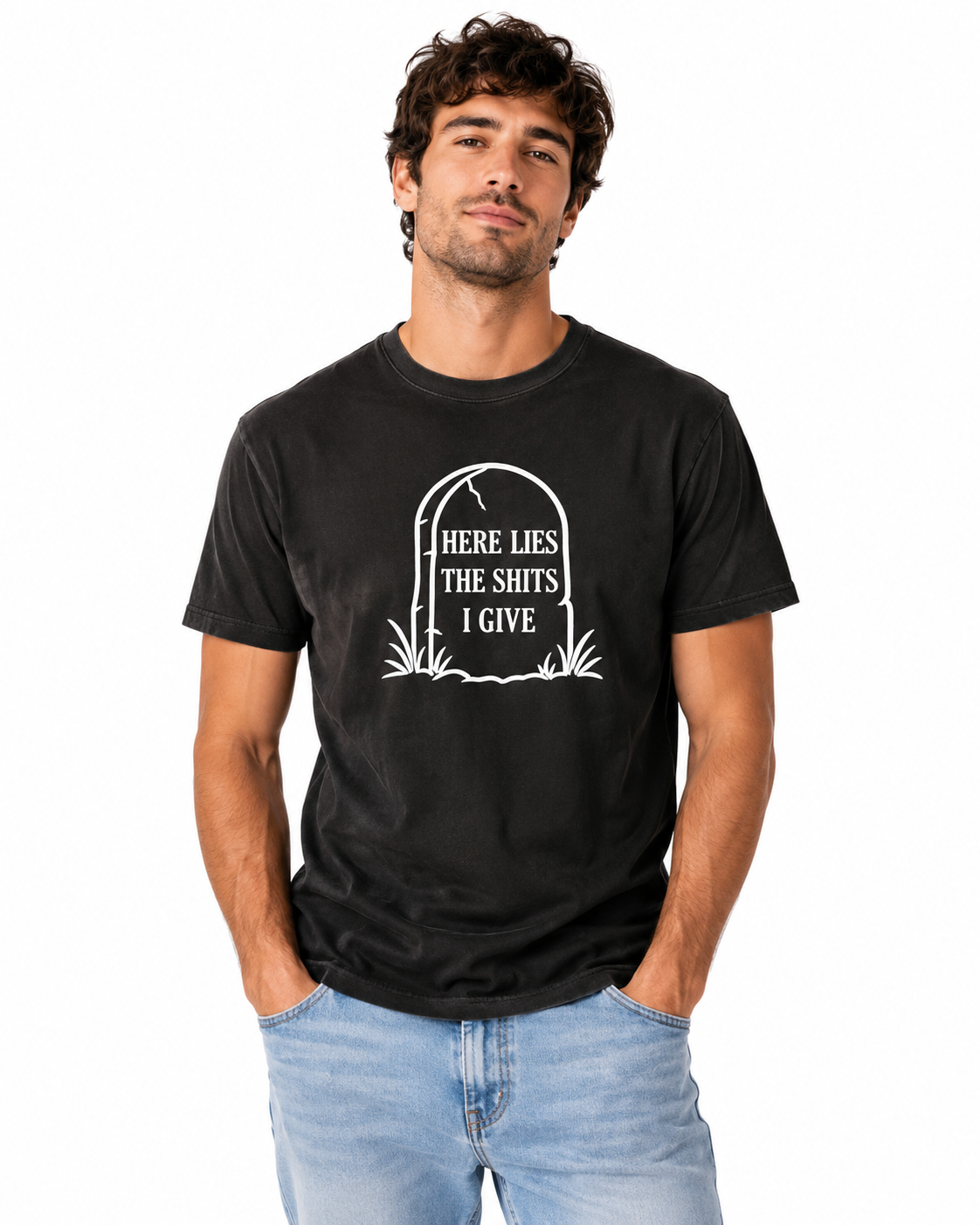 A man wearing a black graphic t-shirt with a white tombstone design that reads 'HERE LIES THE SHITS I GIVE paired with light blue jeans, against a plain white background.