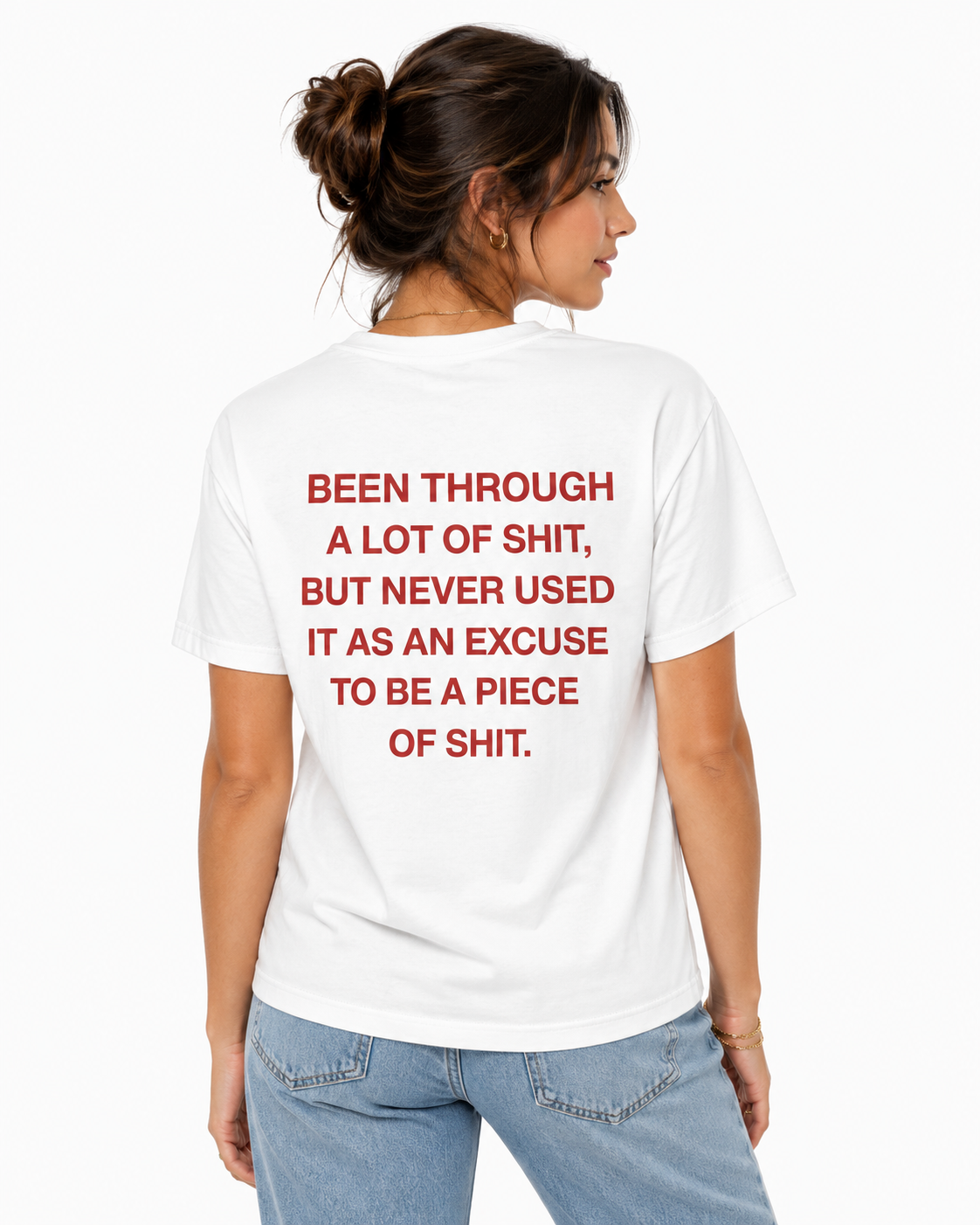A woman shown from the back wearing a white graphic T-shirt with bold red text that reads, 'BEEN THROUGH A LOT OF SHIT, BUT NEVER USED IT AS AN EXCUSE TO BE A PIECE OF SHIT.' She is also wearing light blue jeans against a clean white studio background.