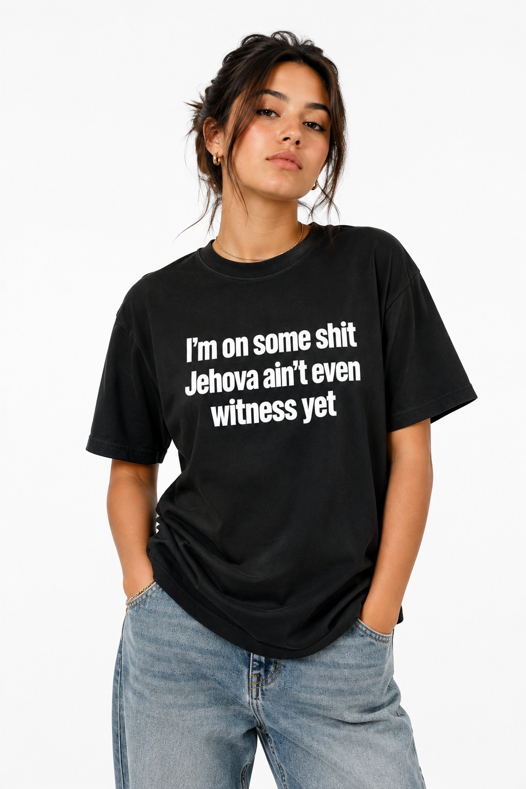 A woman wearing a black graphic t-shirt with white text that reads 'I'm on some shit Jehovah ain't even witness yet', paired with light wash blue jeans, against a plain white background.